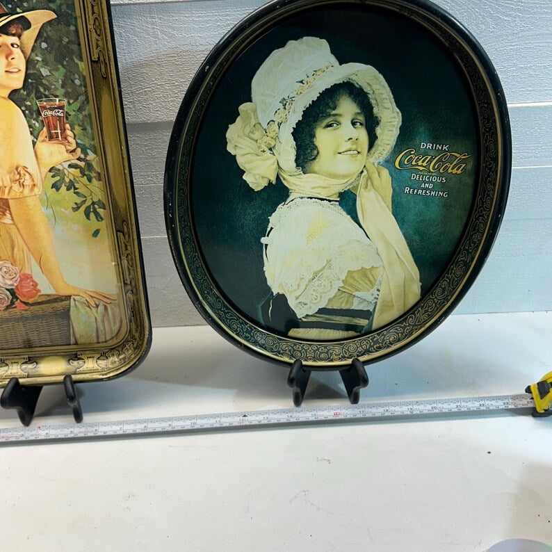 May include: Two vintage Coca-Cola serving trays. One is rectangular with a woman holding a glass of Coca-Cola. The other is oval, featuring a woman in a white hat and the Coca-Cola logo with the text "Delicious and Refreshing."