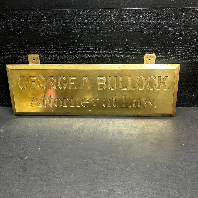 May include: A rectangular brass sign with the engraved text "GEORGE A. BULLOCK. Attorney at Law." The sign has two small holes at the top for mounting. The sign is a golden color.