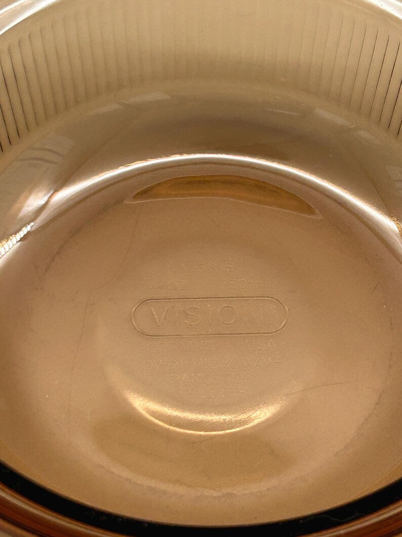 May include: A clear, amber-colored Vision brand glass bowl. The bowl has a rounded shape with a slightly flared rim and a smooth, reflective surface. The word "VISION" is embossed on the bottom.