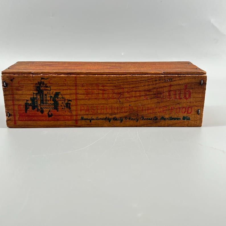 May include: A rectangular wooden box with a reddish-brown finish. The box features printed text and a graphic of a building in black and red. The text includes the words "PASTEURIZED CHEESE FOOD" and "Manufactured by Kraft & Dairy Cheese Co. Maritowoc, Wis."