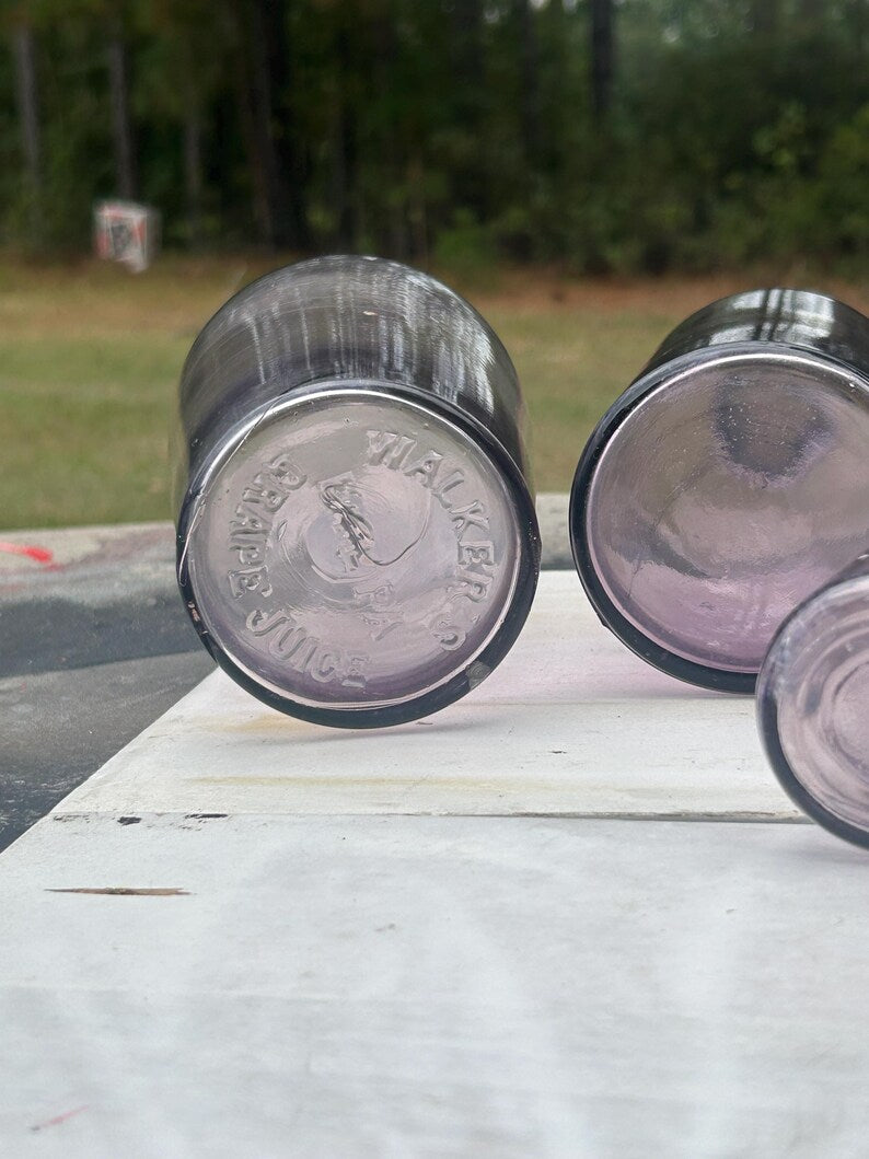May include: Close-up of the bottom of several vintage purple glass bottles. The text "WALKER'S GRAPE JUICE" is embossed on the base of one bottle. The bottles are arranged on a white surface, with a blurred green background.