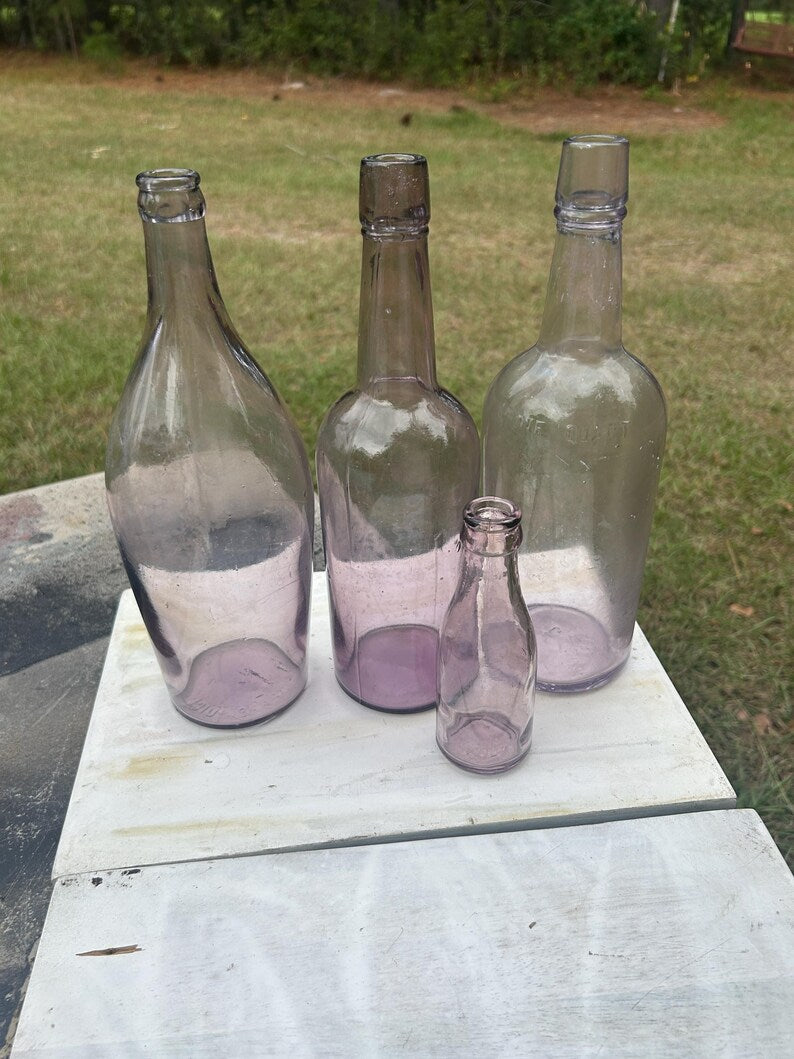May include: Four antique glass bottles in varying sizes and shapes. The bottles are a light purple color and appear to be empty. They are arranged on a white surface, with a green background.