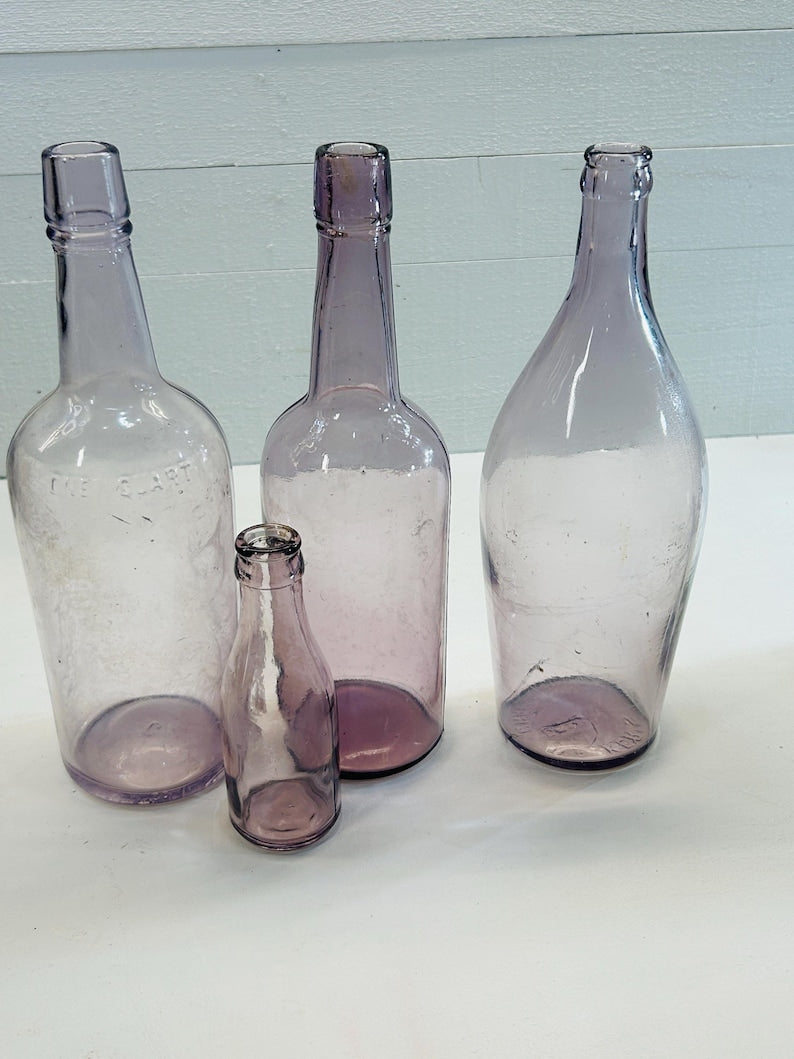 May include: Four antique, clear glass bottles in varying sizes and shapes. The bottles are a soft purple hue, with visible imperfections and signs of age. One bottle has the text "THE CLART" embossed on its side. The bottles are arranged on a white surface.