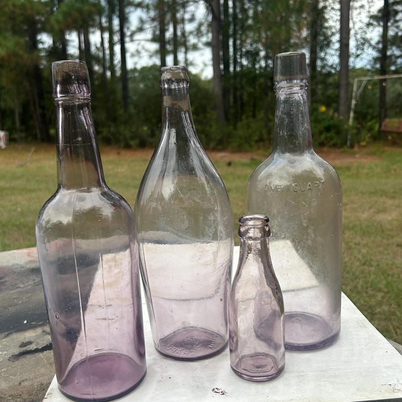 May include: Four antique glass bottles of varying sizes and shapes. The bottles are a light purple color and appear to be empty. The bottles are arranged on a white surface with a blurred background of trees.