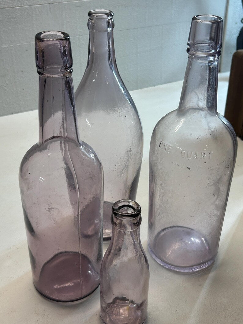 May include: Four antique, clear glass bottles in varying sizes. The bottles have a light purple hue and are arranged on a white surface. One bottle is labeled "ONE QUART". The bottles are empty and have a vintage aesthetic.
