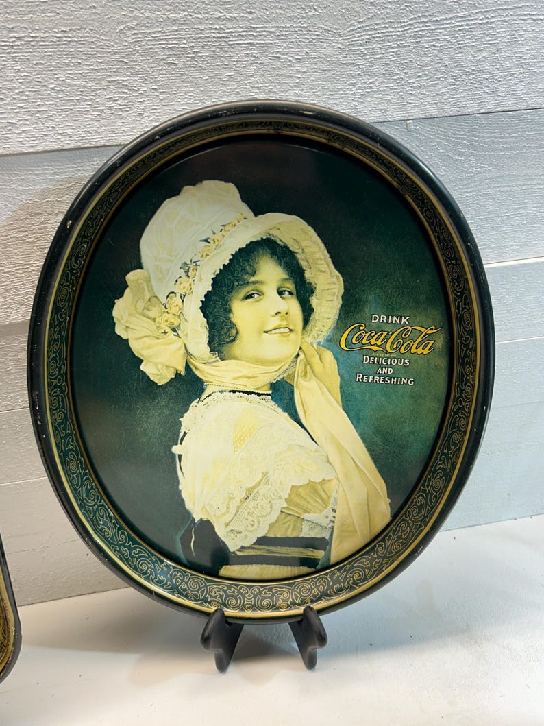 May include: Vintage Coca-Cola serving tray featuring a woman in a white hat and dress. The tray has a dark green background with the words "Drink Coca-Cola Delicious and Refreshing". The tray has a black and gold decorative border.