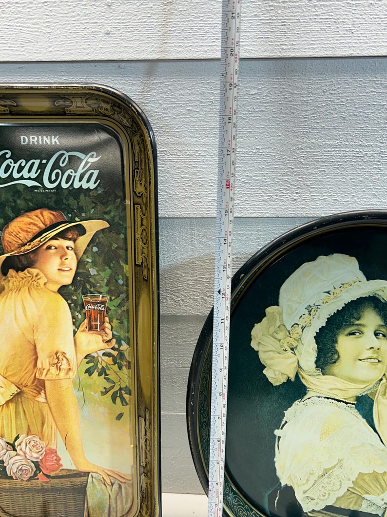 May include: Two vintage Coca-Cola serving trays. One rectangular tray features a woman in a yellow dress holding a glass of Coca-Cola. The other is a round tray with a woman in a white hat. The trays are approximately 19 inches tall.