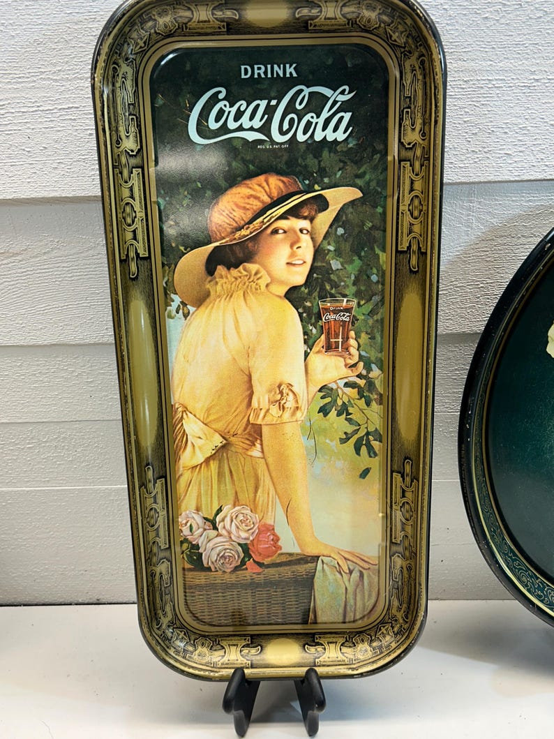 May include: Vintage Coca-Cola serving tray with a woman in a yellow dress and hat holding a glass of Coca-Cola. The tray has a gold and black ornate border and the words "DRINK Coca-Cola" at the top.