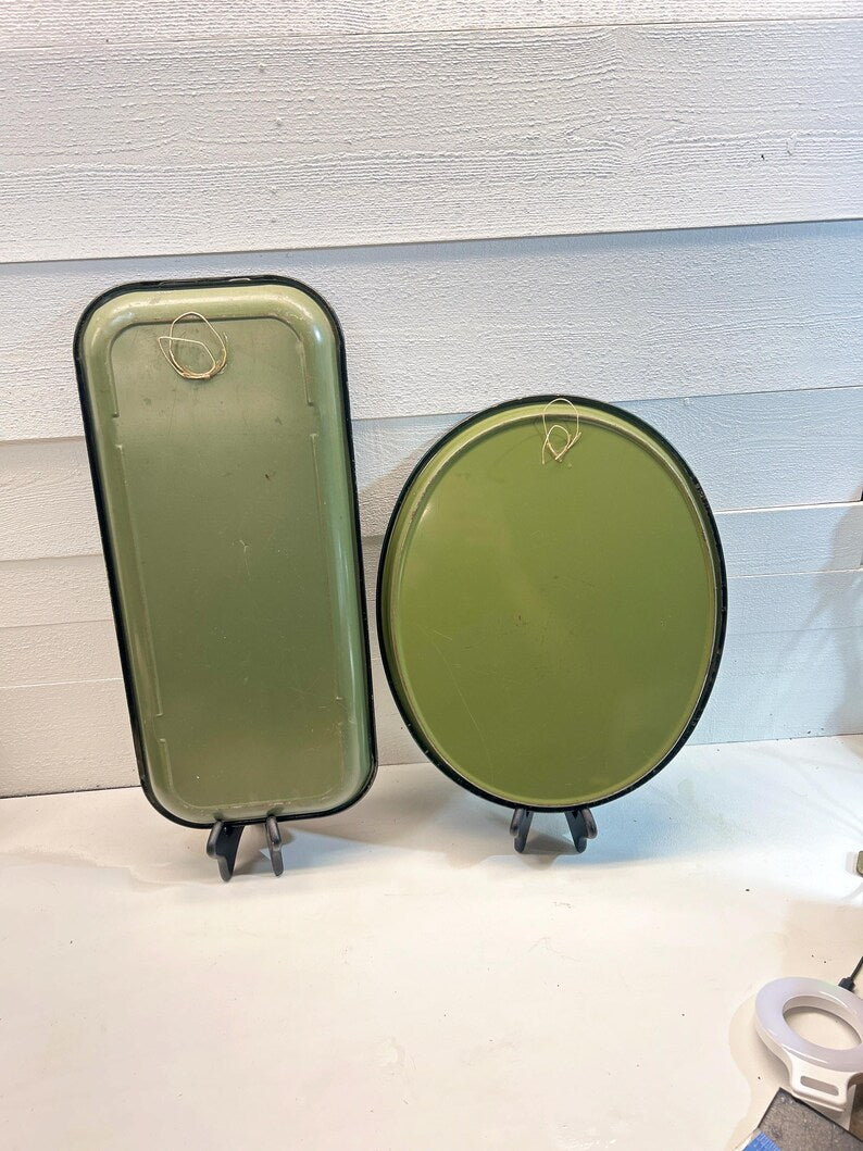 May include: Two olive green metal trays, one rectangular and one round, displayed on black stands. Both trays have a black trim and a wire loop for hanging. The trays are set against a white background.