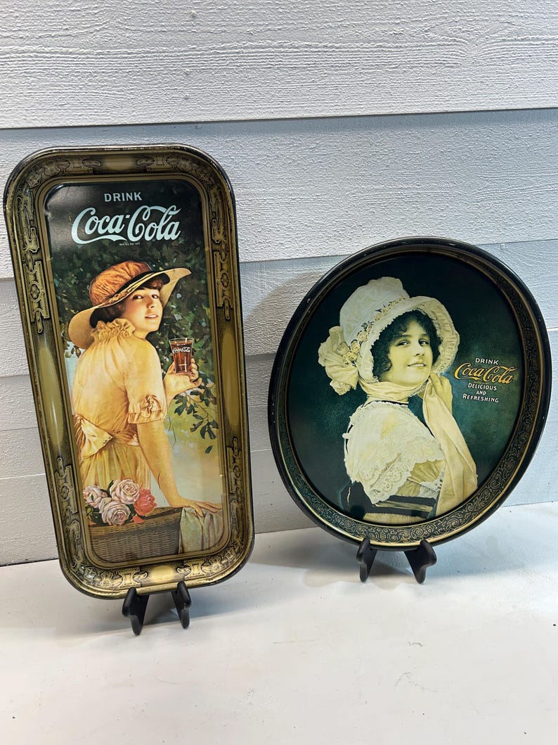May include: Two vintage Coca-Cola serving trays. One is rectangular with a woman in a yellow dress and hat, holding a glass of Coca-Cola. The other is round with a woman in a white hat and dress. Both trays have the Coca-Cola logo.