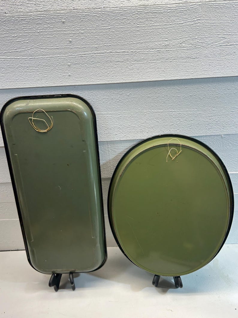 May include: Two olive green metal trays, one rectangular and one round, with black trim. Each tray has a gold wire loop for hanging. The trays are displayed on small black stands against a white wood-paneled background.