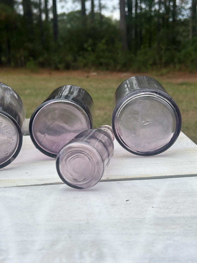 May include: Four antique, lavender-colored glass bottles are arranged on a white surface. The bottles vary in size and shape, with rounded and cylindrical forms. The glass has a slight iridescence, and the bottoms of the bottles are visible.