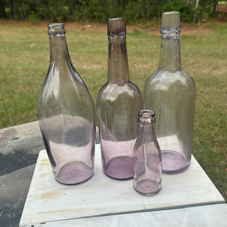 May include: Four antique glass bottles of varying sizes and shapes, in shades of purple and clear. The bottles are arranged on a white surface, with a grassy background. The bottles appear to be vintage, with a classic design.