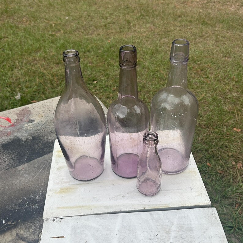 May include: Four antique glass bottles of varying sizes and shapes. The bottles are a light purple color and appear to be empty. They are arranged on a white surface, with a green grassy background.
