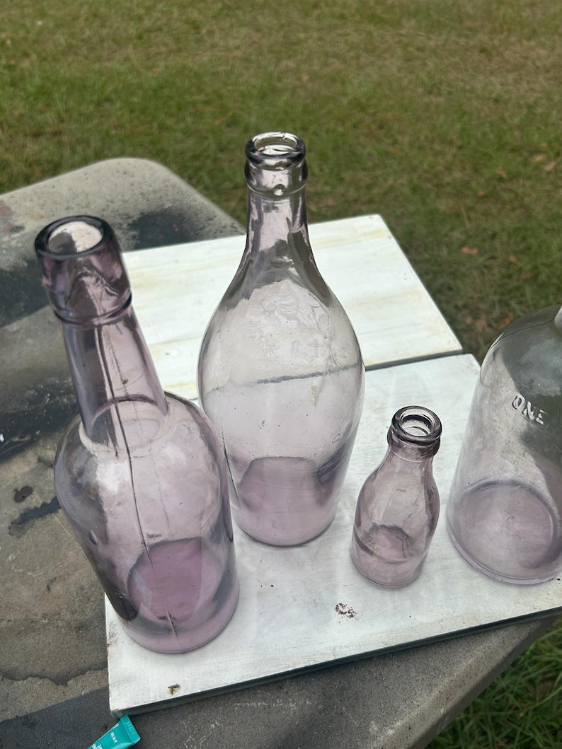May include: Four antique glass bottles in varying sizes and shapes. The bottles are a light purple color and sit on a white surface. The bottles are empty and have a vintage aesthetic.