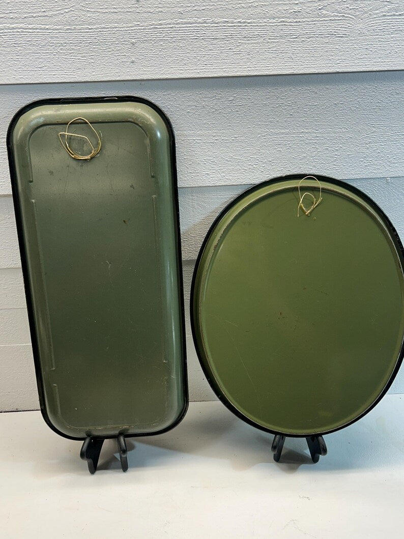 May include: Two vintage metal trays, one rectangular and one round, both in olive green with black trim. Each tray has a gold wire loop for hanging. The trays are displayed on black stands against a white background.