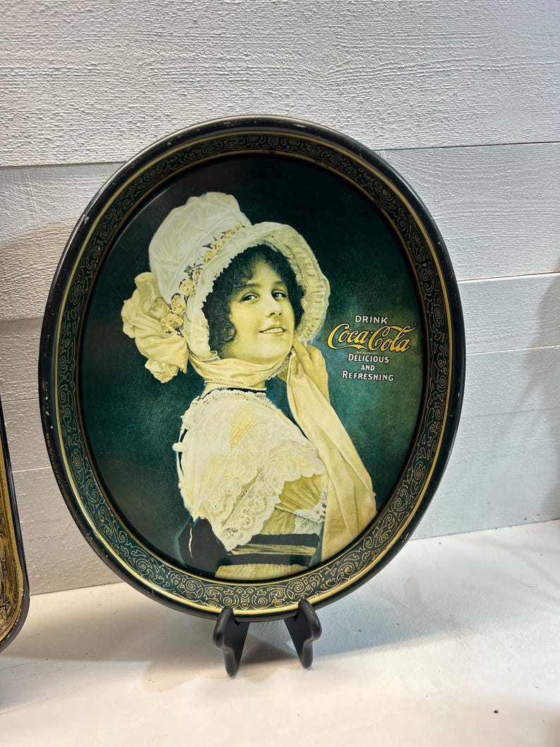May include: Vintage Coca-Cola serving tray featuring a smiling woman in a white hat and dress. The tray has a dark green background with the Coca-Cola logo and the words "Delicious and Refreshing". The tray has a black border with a decorative design.