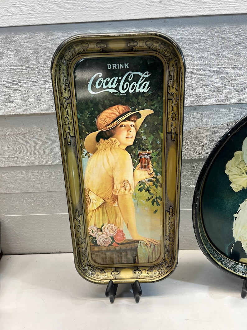 May include: Vintage Coca-Cola serving tray with a woman in a yellow dress and hat holding a glass of Coca-Cola. The tray has a gold-colored frame and the words "DRINK Coca-Cola" at the top. The woman is standing in a garden setting.