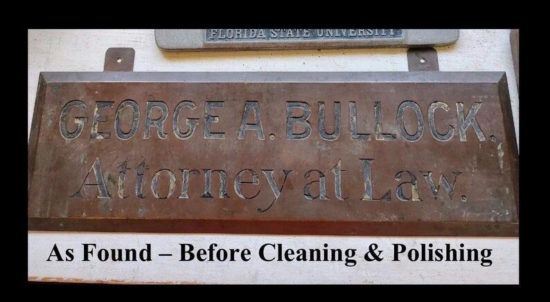 May include: A weathered bronze sign with the engraved text "GEORGE A. BULLOCK, Attorney at Law." The sign has two mounting holes and reads "As Found - Before Cleaning & Polishing" at the bottom. Another sign is visible in the background.