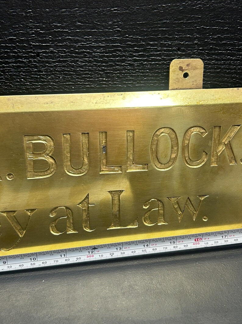 May include: A rectangular brass sign with the engraved text "BULLOCK" and "at Law." The sign has a small mounting bracket at the top and a ruler along the bottom. The sign is a golden color.