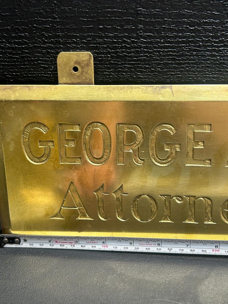 May include: A rectangular brass sign with the name "GEORGE" and the word "Attorney" engraved in capital letters. The sign has a small rectangular bracket at the top for hanging. A measuring tape is at the bottom.