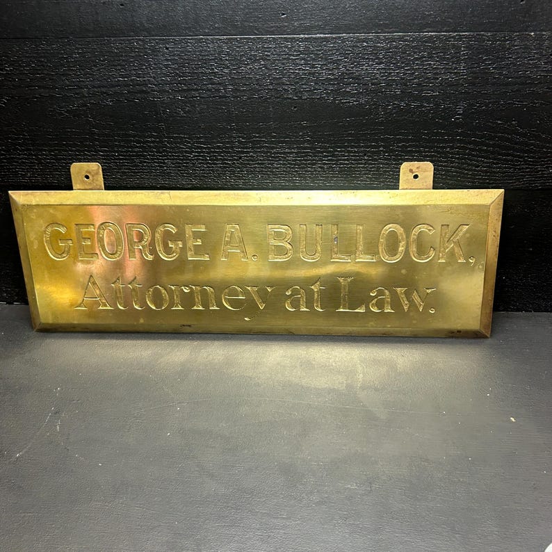 May include: A rectangular brass sign with the engraved text "GEORGE A. BULLOCK, Attorney at Law." The sign has two mounting holes at the top and is set against a dark background.