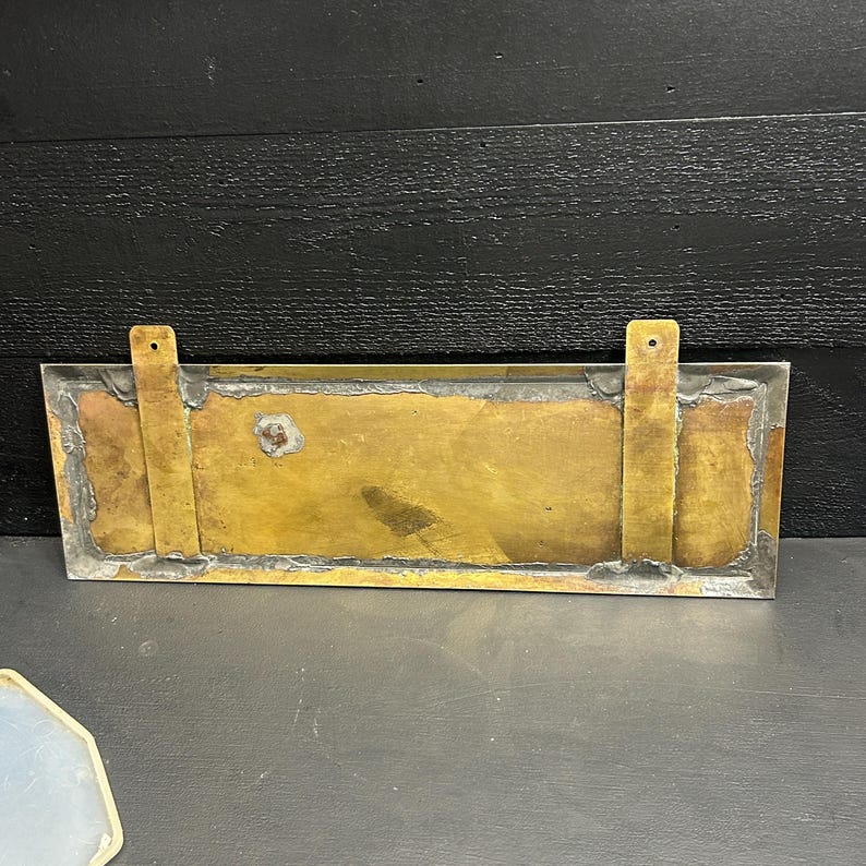 May include: A rectangular brass plate with two vertical mounting tabs. The plate has a tarnished, aged appearance with visible solder seams and a slightly irregular shape. The brass has a warm, golden hue, and the edges are framed with a silver-colored metal.
