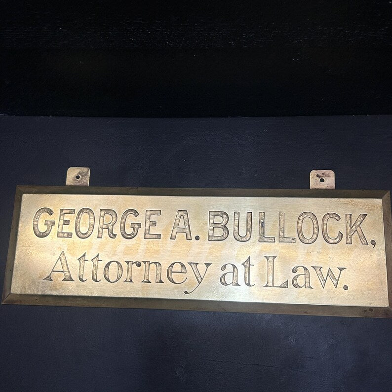 May include: A rectangular brass sign with the engraved text "GEORGE A. BULLOCK, Attorney at Law." The sign has a dark background and two mounting holes at the top.