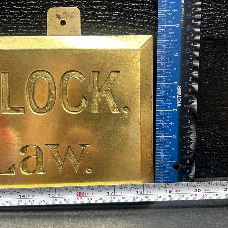 May include: A rectangular brass sign with the word "LOCK." and "LAW." engraved in bold letters. A blue ruler is placed next to the sign. The sign has a small hole at the top for hanging.