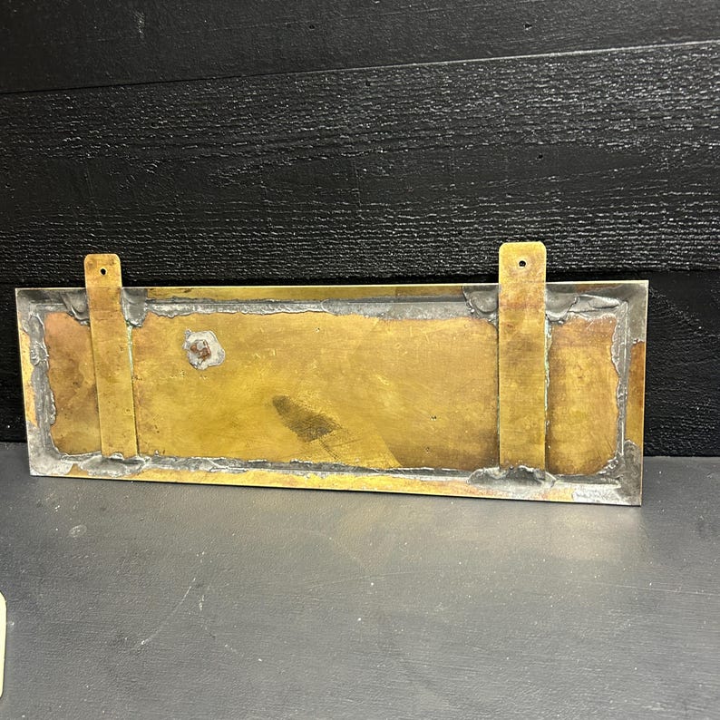 May include: A rectangular brass mailbox plate with two vertical mounting brackets. The plate has a tarnished gold finish with a silver border and is mounted on a dark surface.