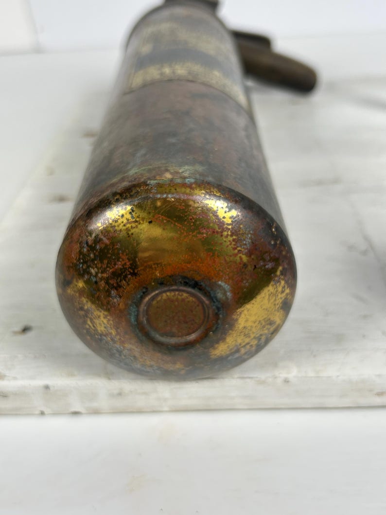 May include: Antique brass soda siphon with a tarnished finish. The bottom of the siphon has a gold and brown patina. The handle and top are a darker brass color. The siphon is resting on a white surface.