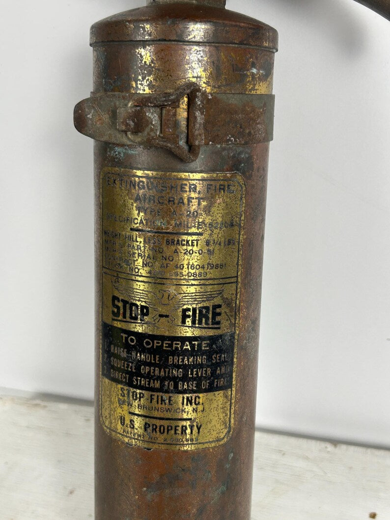 May include: Vintage aircraft fire extinguisher with a copper-colored body and gold labels. The labels include text such as "STOP - FIRE" and instructions for use. The extinguisher has a metal clasp and handle.