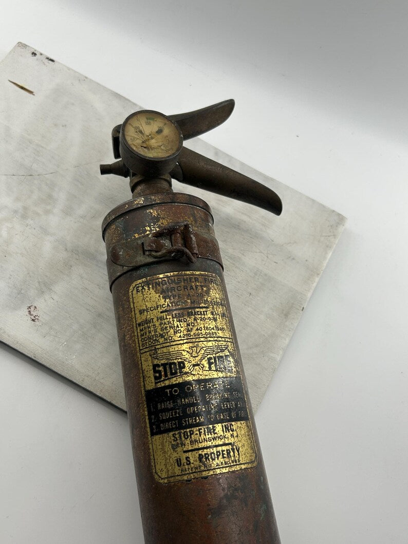 May include: Vintage aircraft fire extinguisher with a brass finish. The extinguisher has a gauge, a handle, and a label that reads "STOP-FIRE". The label also includes operating instructions and the text "U.S. PROPERTY".