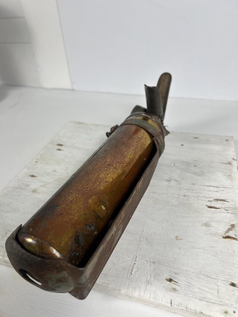 May include: An antique brass and metal oil lamp with a cylindrical body and a curved spout. The lamp has a dark, aged patina, suggesting it is vintage. The lamp is set in a metal frame and sits on a white wooden surface.