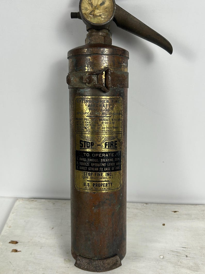 May include: Vintage Stop-Fire aircraft fire extinguisher. The cylindrical metal body has a bronze finish with a gauge at the top and a lever handle. The label reads "Stop-Fire" and "U.S. Property."