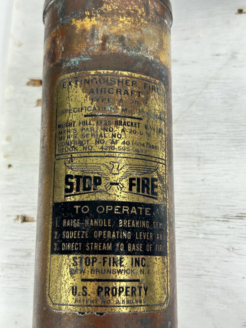 May include: Vintage aircraft fire extinguisher with a weathered, metallic finish. The label reads "STOP-FIRE" and includes instructions for use. The extinguisher is marked "U.S. PROPERTY".