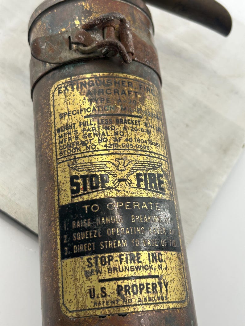 May include: Vintage Stop-Fire aircraft fire extinguisher. The brass-colored cylinder has a label with text including "Stop-Fire Inc." and "U.S. Property." The extinguisher shows signs of age and wear.