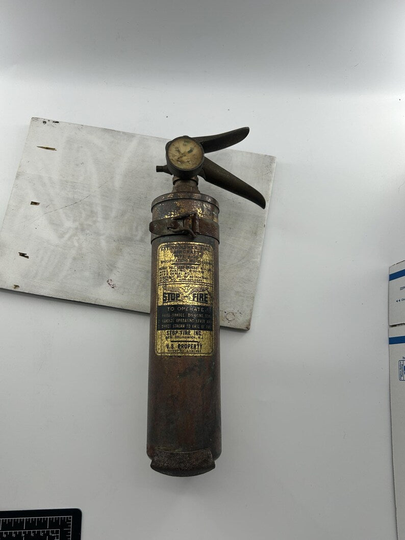 May include: Vintage Stop-Fire Inc. fire extinguisher. The cylindrical body is a dark, aged brown with a gold label. The top features a pressure gauge and a lever handle. The extinguisher is marked "U.S. PROPERTY."