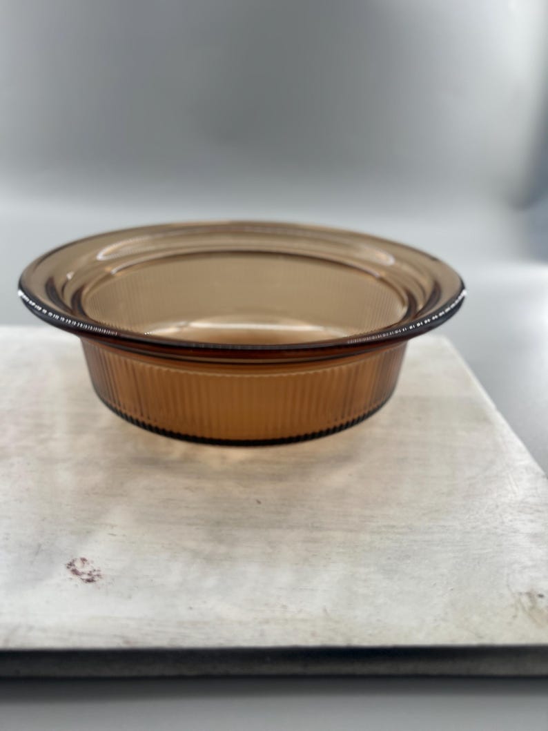 May include: A round, amber-colored glass baking dish with a ribbed exterior. The dish has a slightly flared rim and sits on a light-colored surface. The glass has a warm, transparent quality.