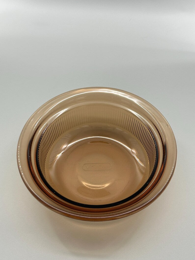May include: Amber-colored glass bowl with a ribbed interior and a smooth exterior. The bowl has a round shape and a slightly flared rim. The brand name is visible on the bottom of the bowl.