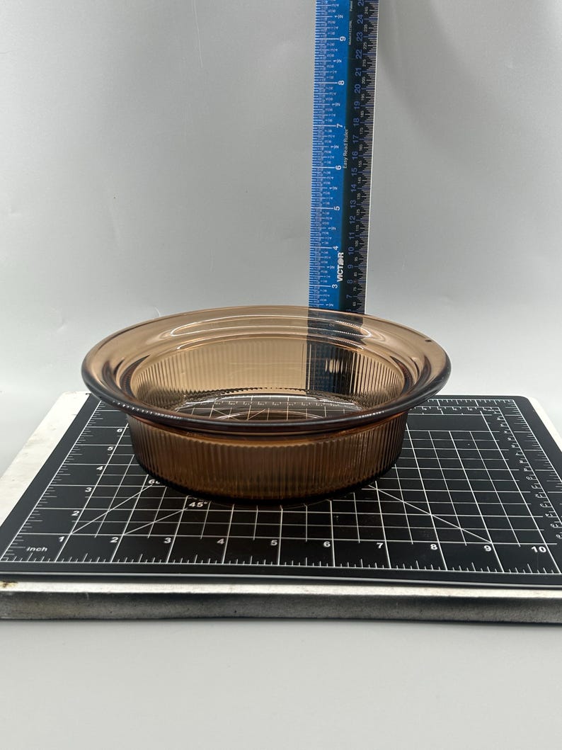 May include: A round, amber-colored glass bowl with a ribbed exterior. The bowl sits on a black and white cutting mat with inch measurements. A blue ruler is in the background.