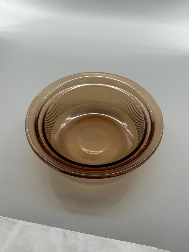 May include: Amber-colored glass bowl with a ribbed interior and a smooth exterior. The bowl is round and nested inside a slightly larger bowl of the same color. The bowls are made of a transparent glass material.