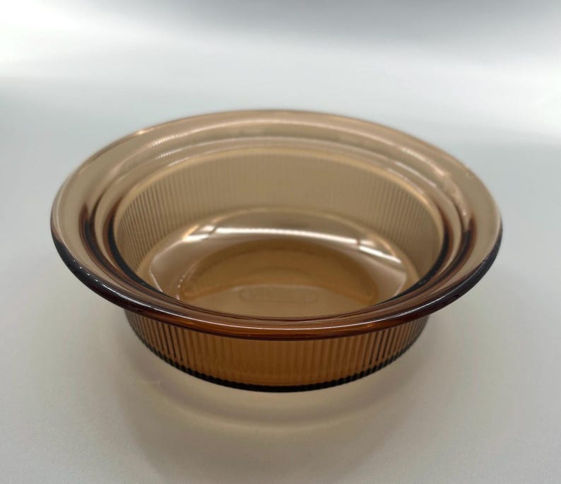 May include: Amber-colored glass bowl with a ribbed exterior and a smooth interior. The bowl has a rounded shape with a slightly flared rim. The glass appears to be vintage or retro in style, suitable for serving or display.