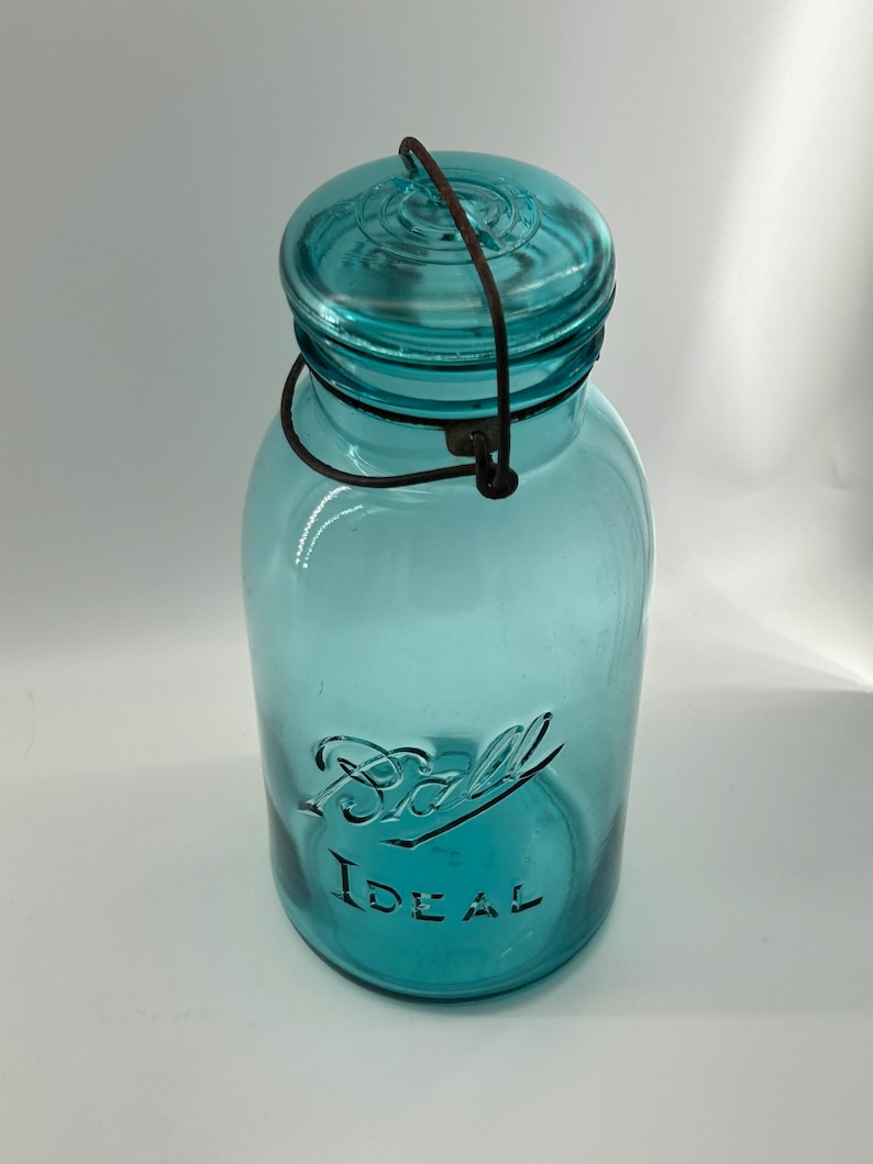 May include: A vintage turquoise Ball Ideal jar with a wire handle and lid. The jar has the embossed text "Ball Ideal" on the front. The jar is made of glass and is likely used for canning or storage.