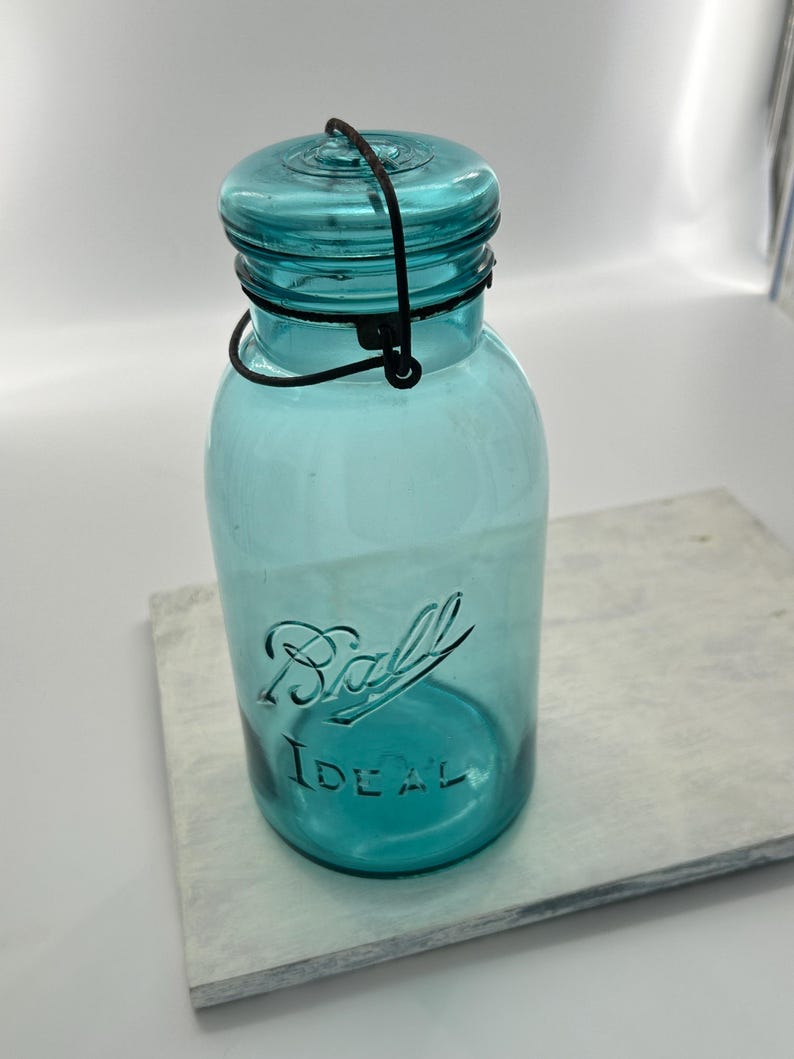 May include: A vintage turquoise Ball Ideal jar with a matching lid and wire handle. The jar has the Ball Ideal logo embossed on the front. The jar is sitting on a white and gray surface.