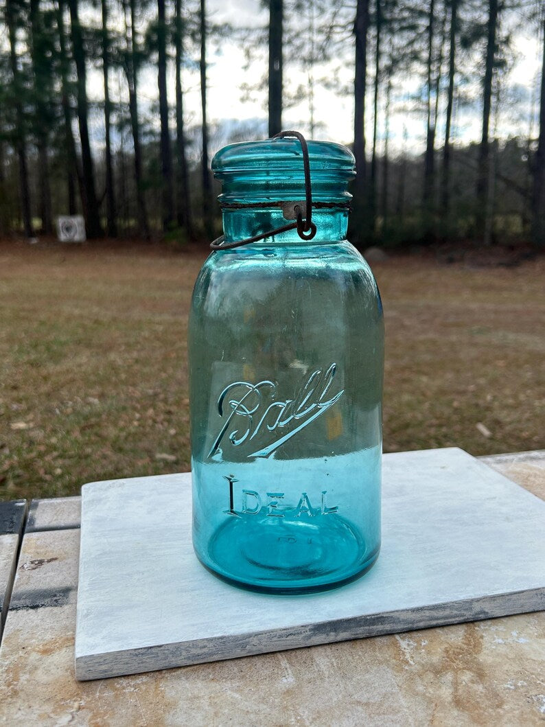 May include: A vintage turquoise Ball Ideal jar with a glass lid and wire closure. The jar has the word "Ball" embossed on the front. The jar is sitting on a white surface.