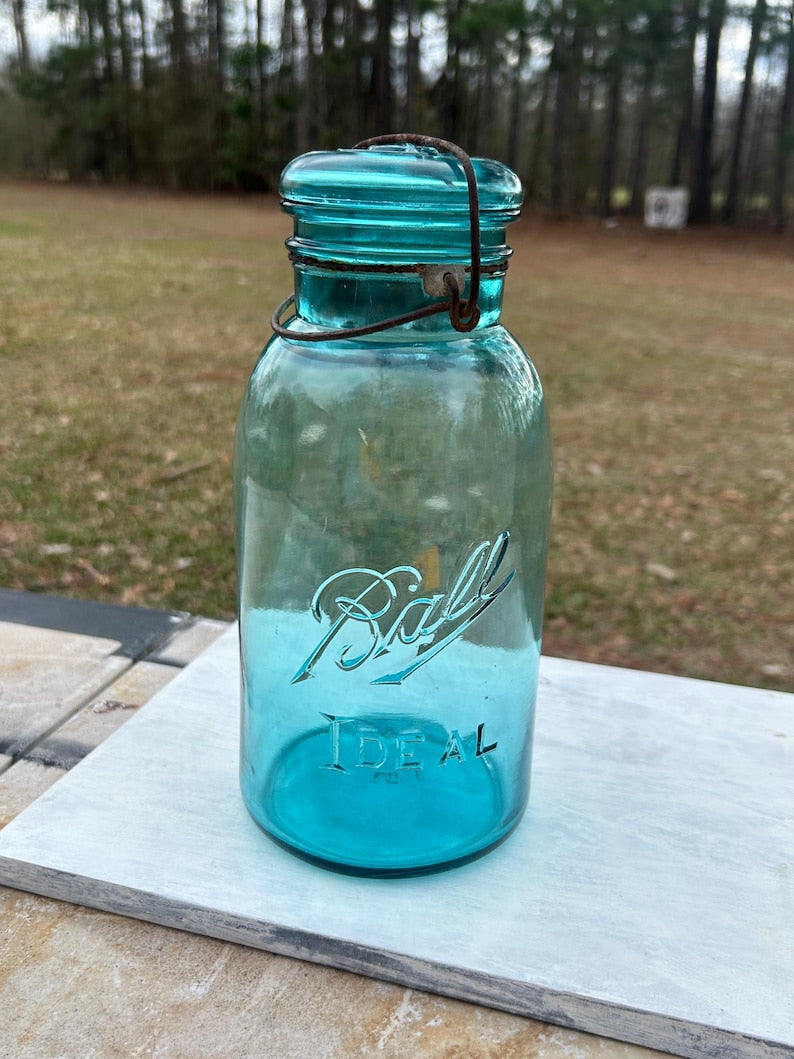 May include: A vintage turquoise Ball Ideal jar with a wire closure. The jar has the word "Ball" in script and "Ideal" embossed on the front. The jar is made of glass and has a metal lid. The jar is sitting on a white surface.