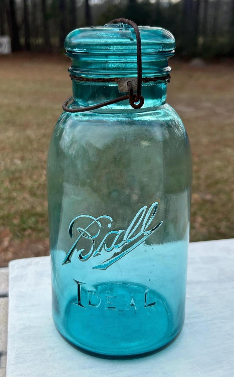 May include: A vintage blue Ball Ideal jar with a wire closure. The jar has the word "Ball" in script and "IDEAL" embossed on the front. The glass has a slight aqua tint, and the jar is likely made of glass.
