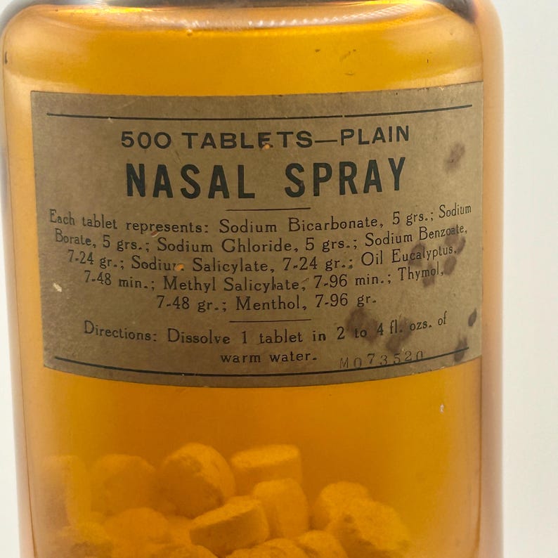 May include: Amber glass bottle labeled "NASAL SPRAY" with "500 TABLETS-PLAIN" printed at the top. The label lists ingredients and directions. The bottle contains yellow tablets. A vintage pharmaceutical item.