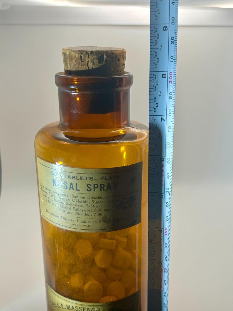 May include: A vintage amber glass bottle with a cork stopper, labeled "NASAL SPRAY." The bottle contains small, yellow tablets. A ruler shows the bottle is approximately 8 inches tall. The label lists ingredients and directions.